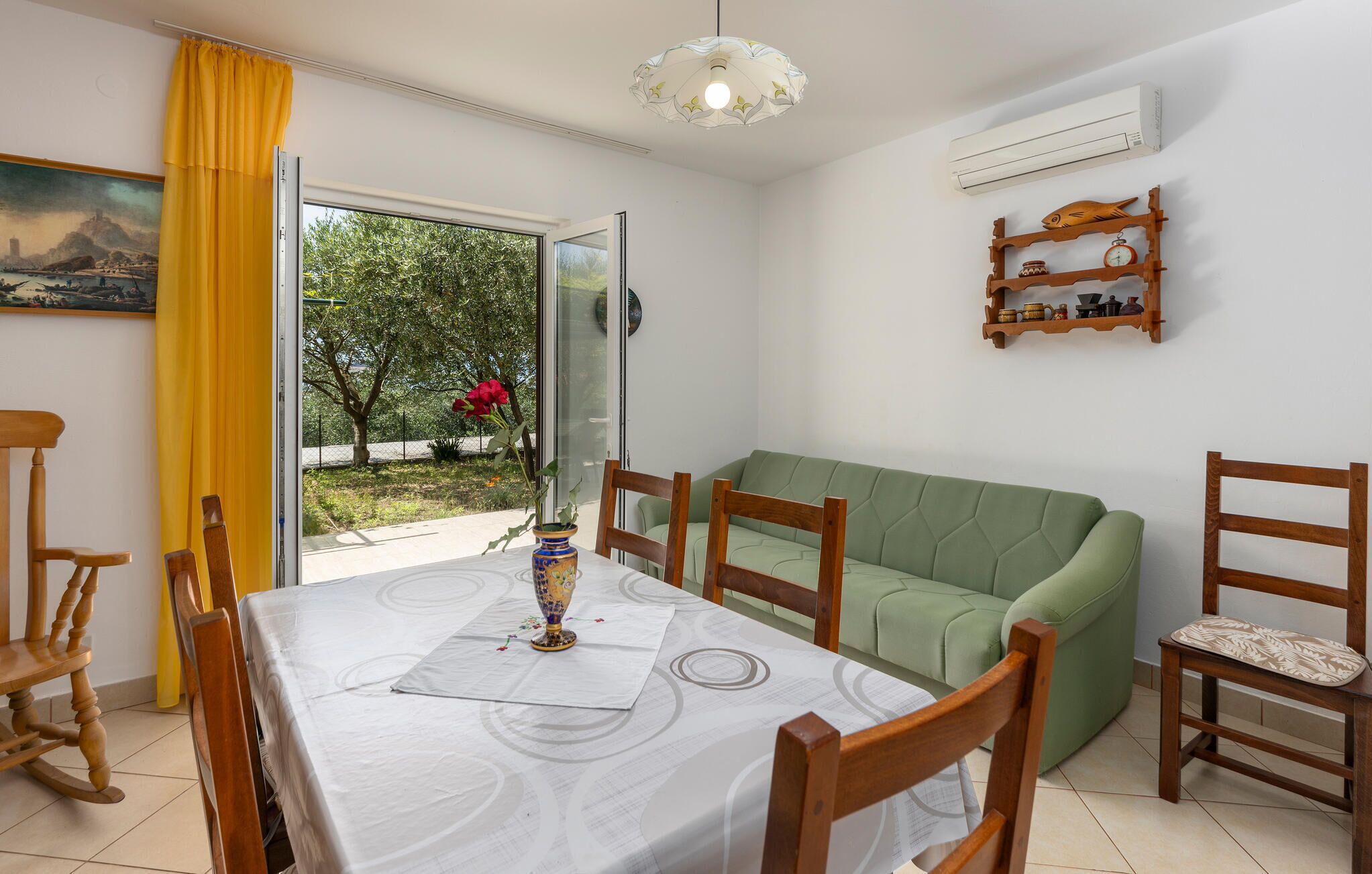A dining area with a table set for four, a green sofa, wooden chairs, and a window with a view of greenery and a red flower.