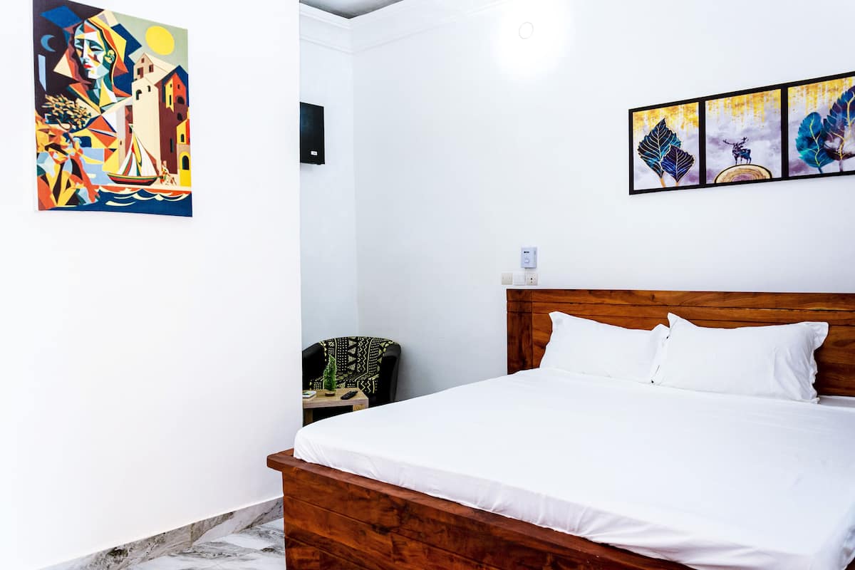 Quarto de hotel com cama de madeira, roupa de cama branca, uma mesinha com uma planta, uma cadeira e obras de arte nas paredes.