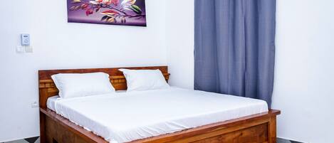 Una cama de madera con ropa de cama blanca, un cuadro floral morado en la pared y una cortina gris.