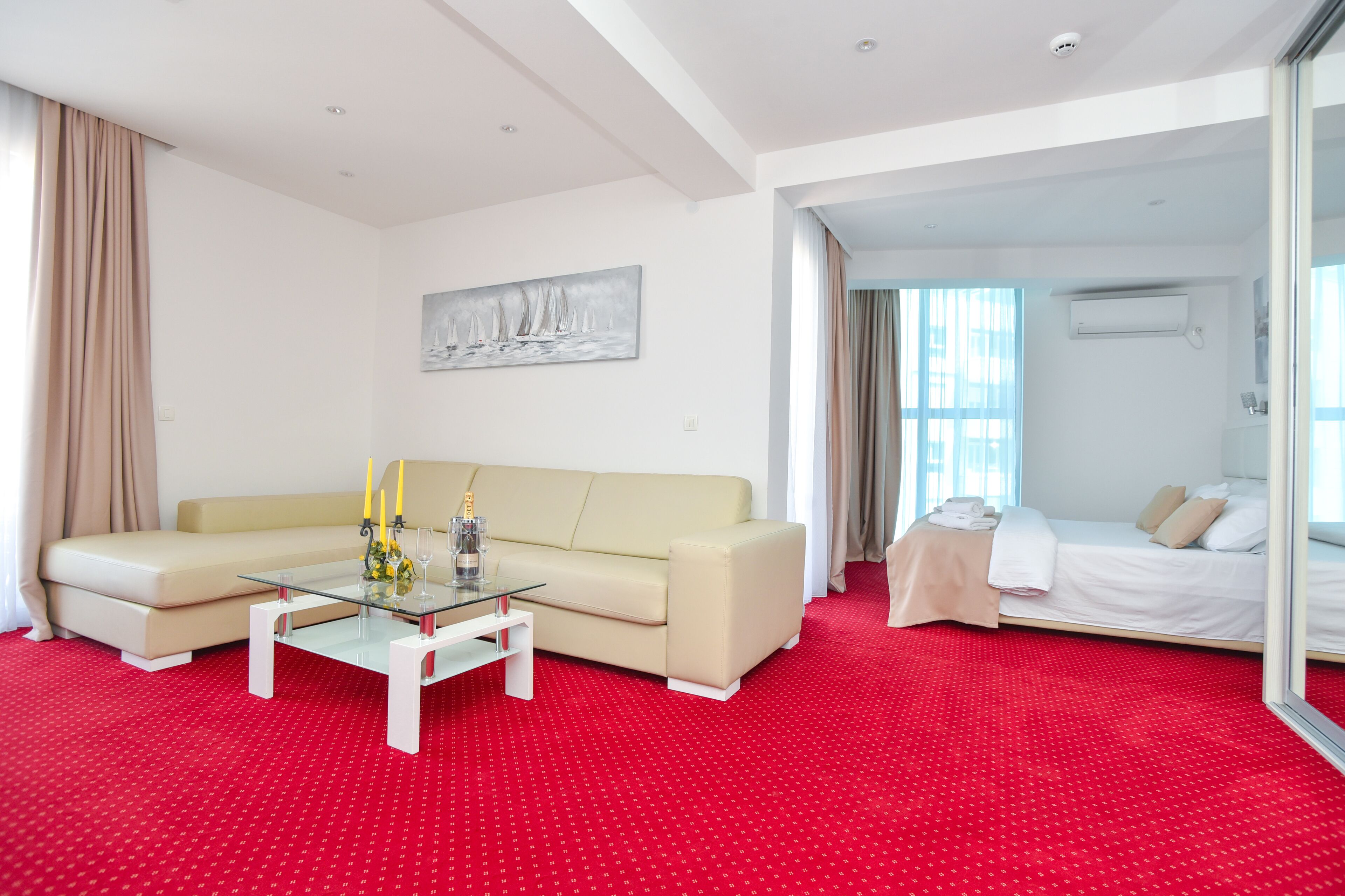 A hotel room with a red carpet, a beige sofa, a glass coffee table with candles, and a bed with a white duvet.