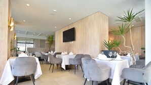 Un comedor moderno con mesas dispuestas para dos, sillas grises tapizadas y un televisor de pantalla plana montado en la pared.