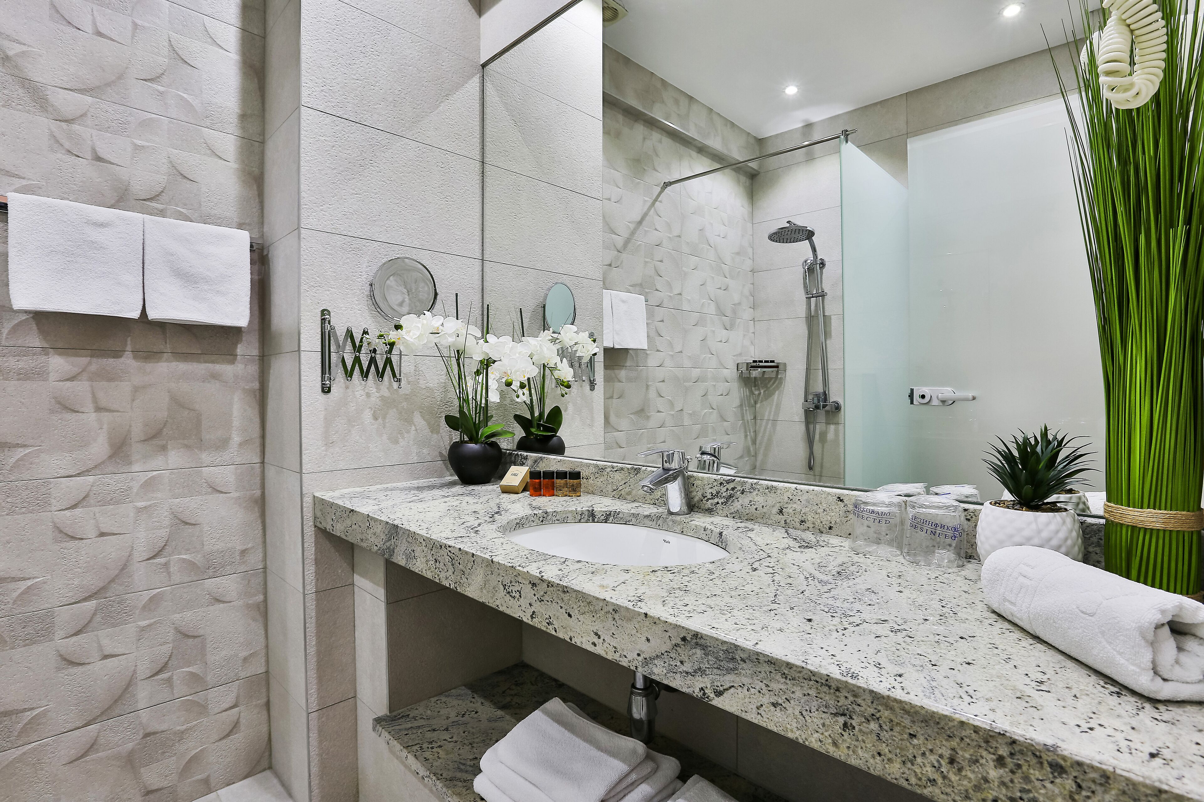 Un baño moderno con encimera de granito, un lavamanos y una ducha. Hay toallas colgadas en la pared y una planta en maceta sobre la encimera.
