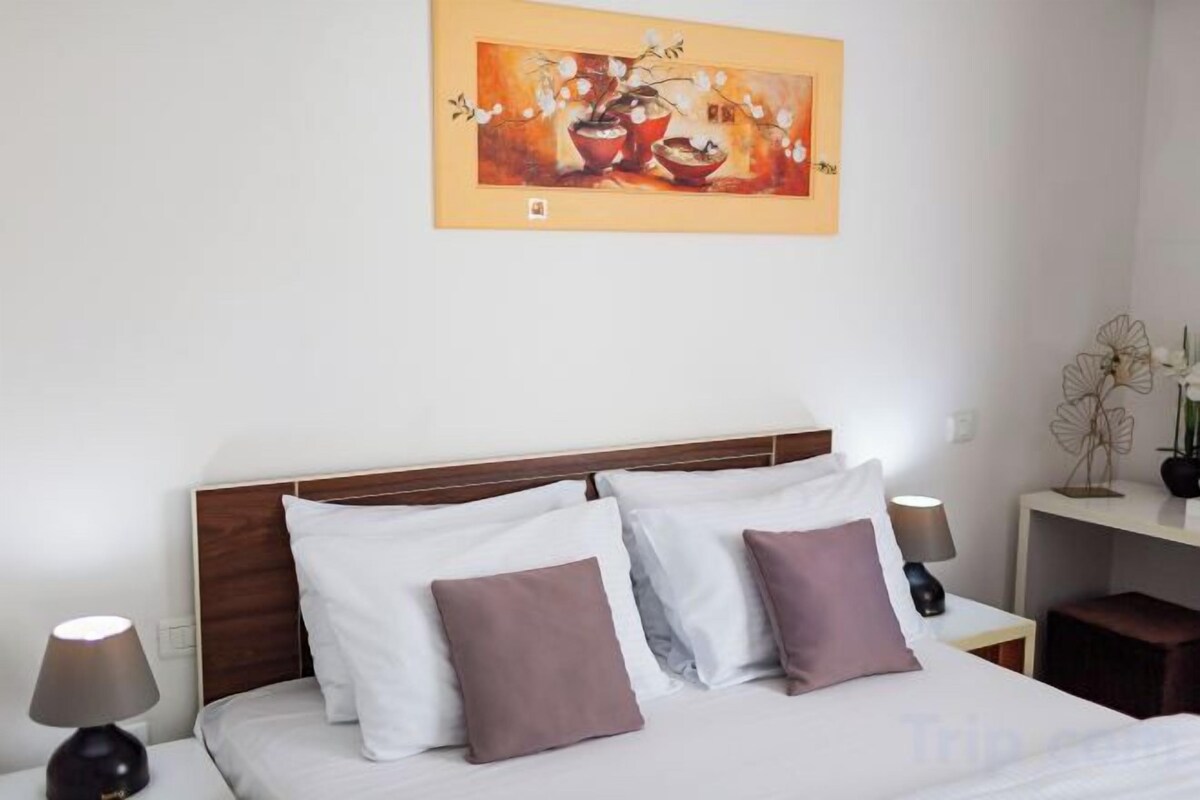 A hotel room with a bed, pillows, a nightstand, a lamp, and a wall-mounted painting.