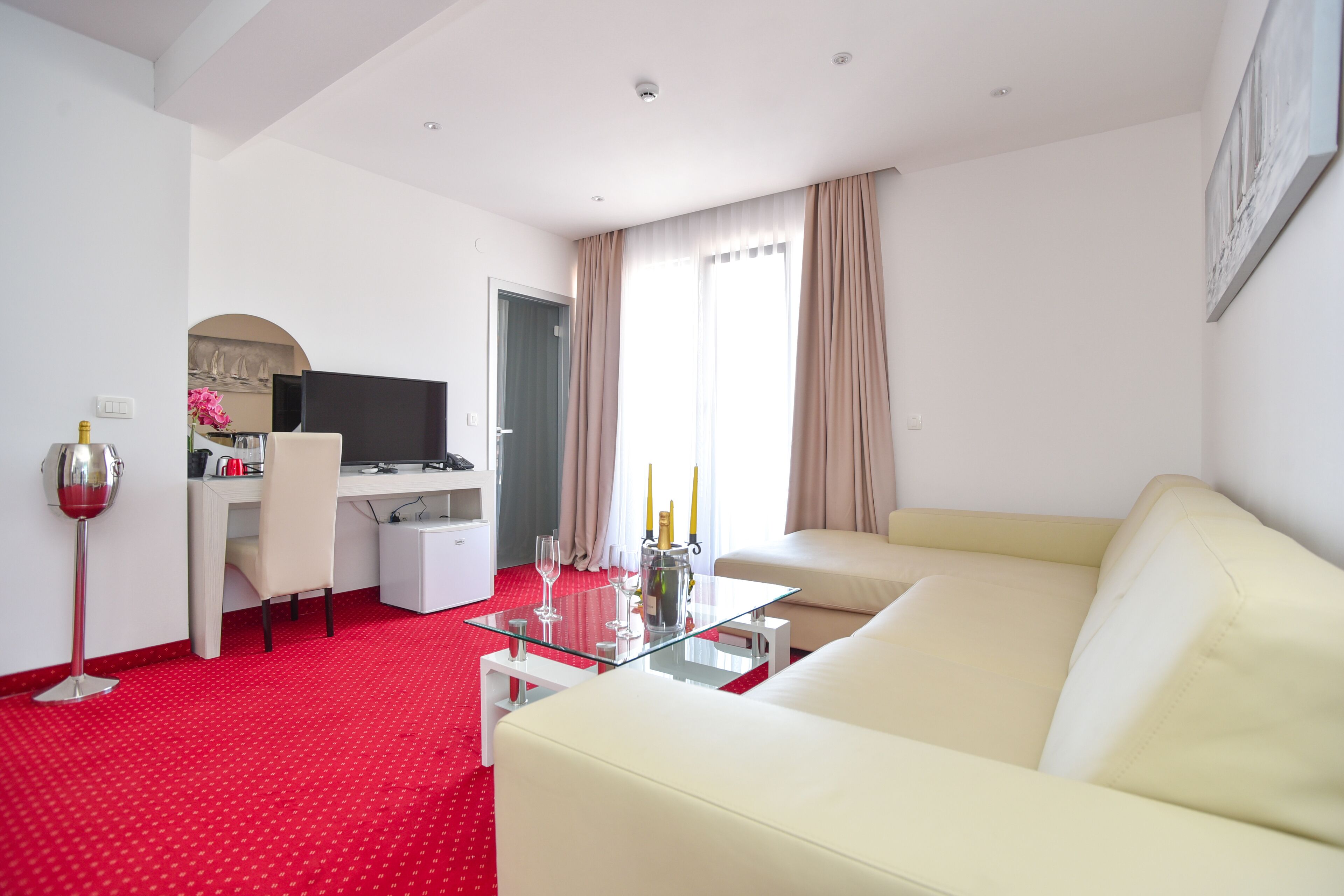 Habitación de hotel con alfombra roja, sofá blanco, mesa de centro de vidrio, escritorio con computadora y televisor.