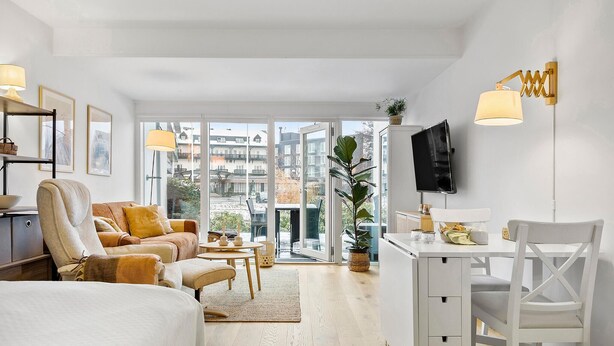 Ein modernes Hotelzimmer mit einem großen Bett, einer Sitzecke, einem Schreibtisch mit Stuhl, einem Fernseher und Blick auf die Stadt durch große Fenster.