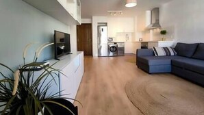 Uma sala de estar moderna com um sofá, uma TV de tela plana e uma cozinha ao fundo.