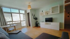 Uma sala de estar moderna com um conjunto de sofá, uma TV sobre um armário branco e uma janela grande com cortinas.