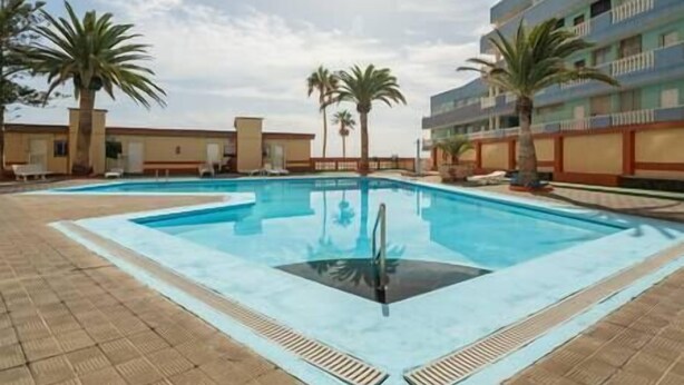 Piscina com deck revestido, cercada por palmeiras e um edifício com varandas.