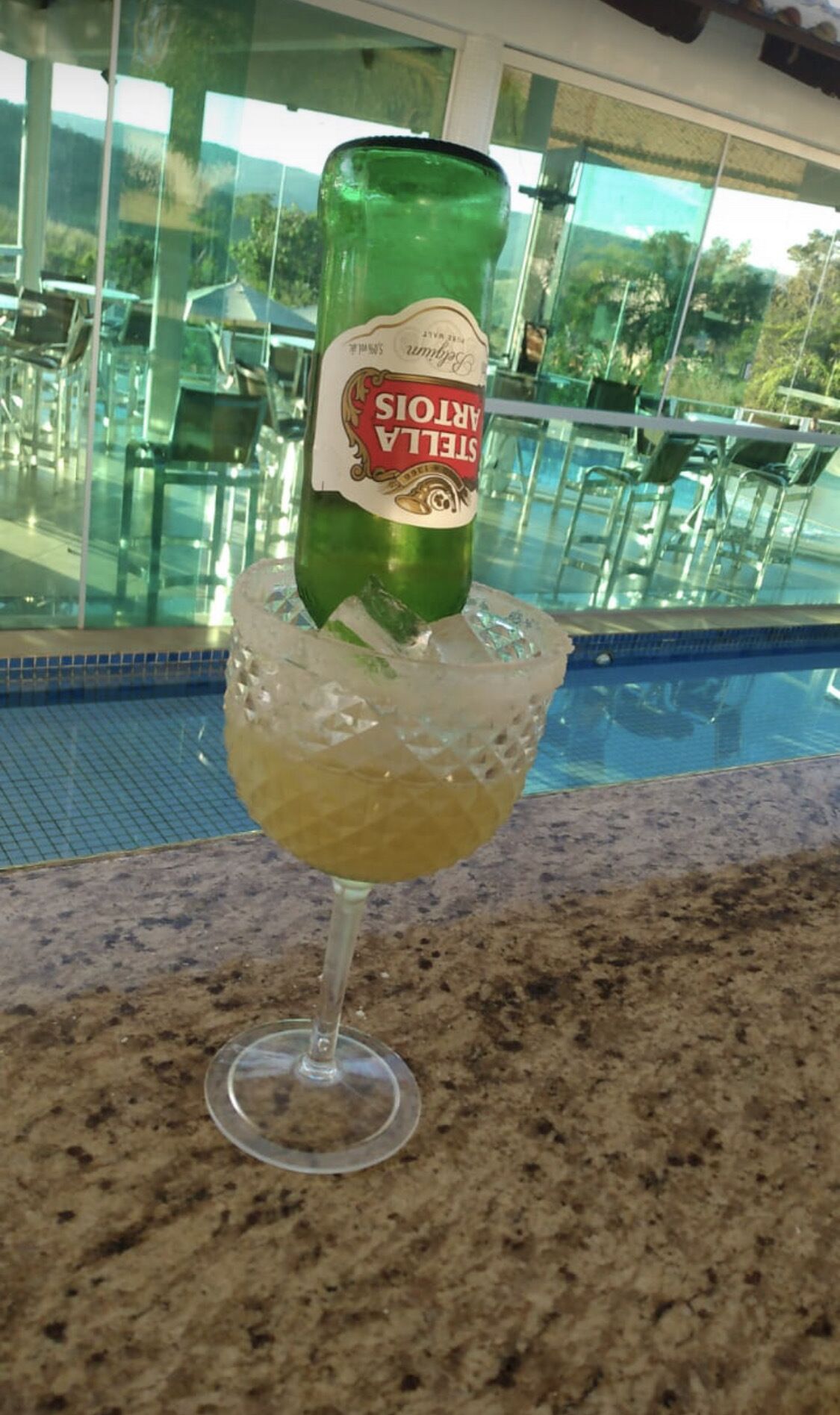 A bottle of Stella Artois beer in a glass with ice cubes, placed on a granite countertop near a pool area with glass chairs and a view of greenery.