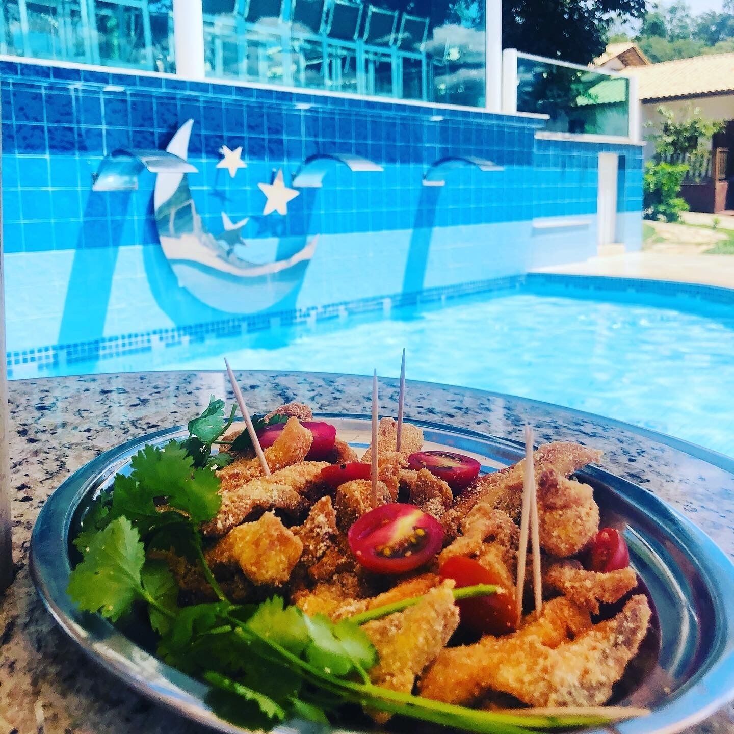 Plato de comida rebozada y frita, adornado con hierbas y verduras, con una piscina al fondo.