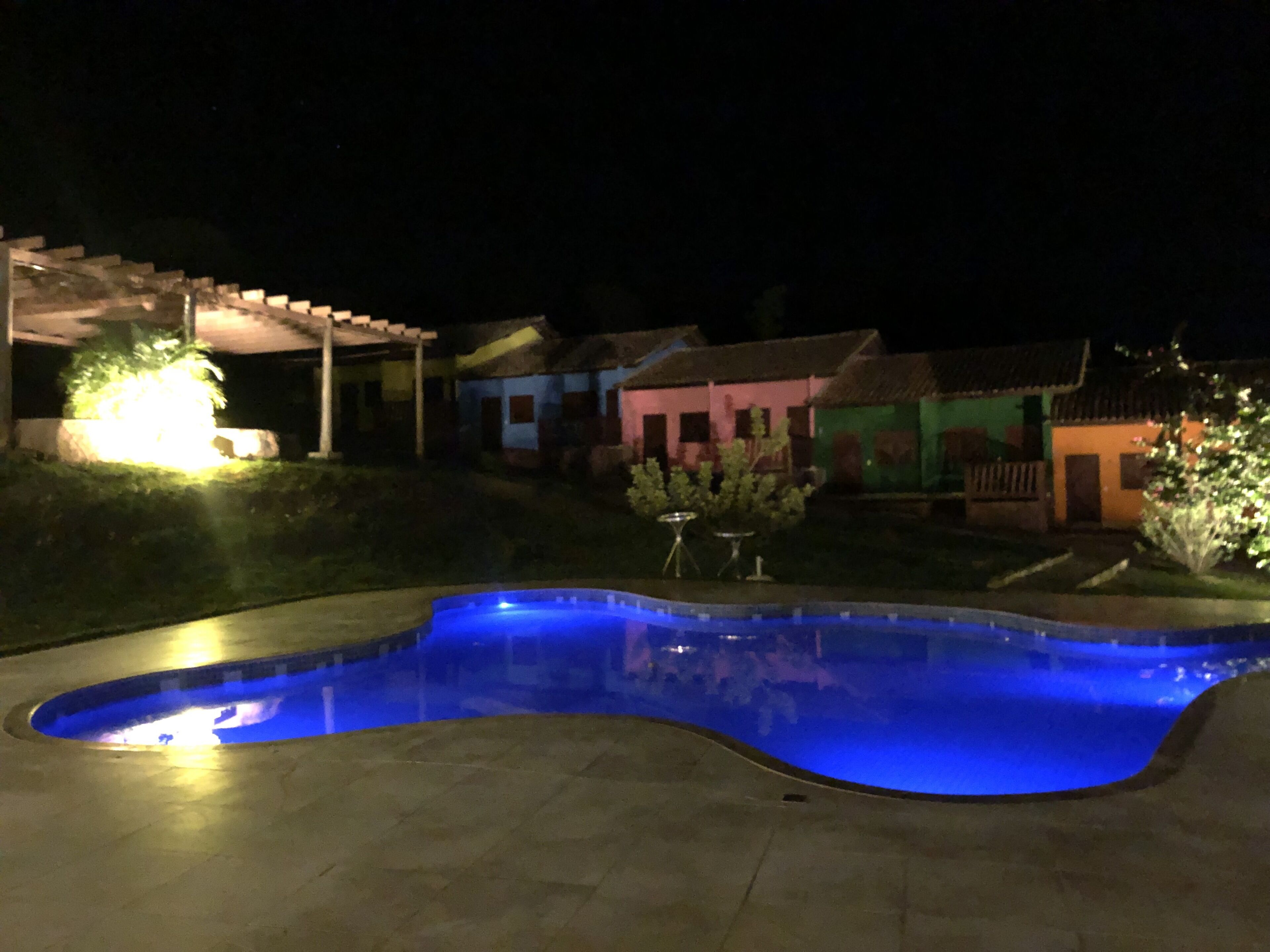 Una piscina de borde curvo, rodeada por una zona pavimentada. Se observan casas con fachadas coloridas al fondo.