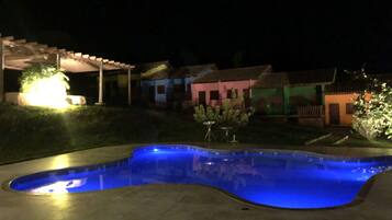 Una piscina de borde curvo, rodeada por una zona pavimentada. Se observan casas con fachadas coloridas al fondo.