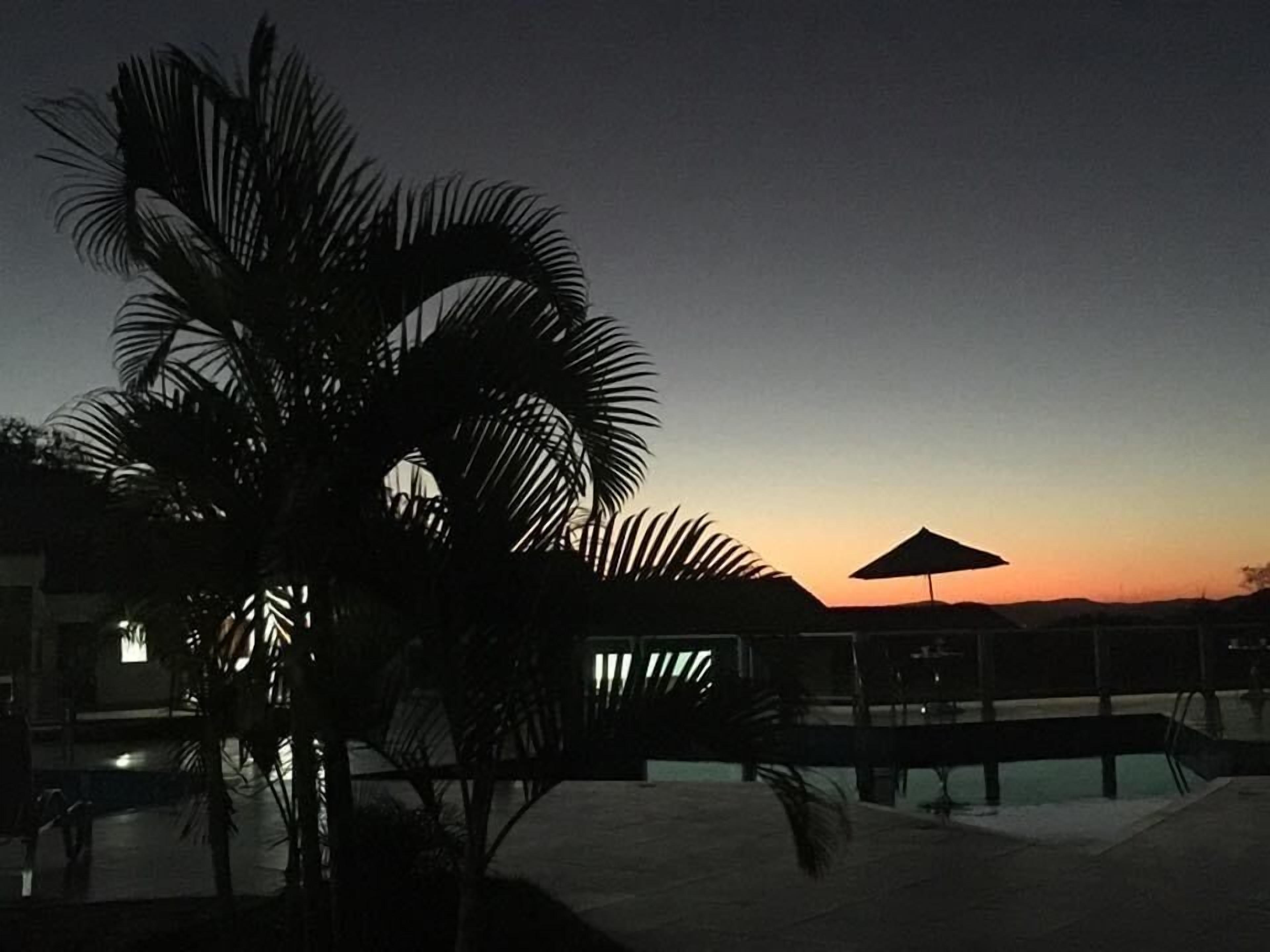 A resort pool area at dusk with palm trees and a sunset in the background.