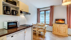 Cocina compacta con horno integrado, cafetera y lavavajillas. Hay una mesa de comedor con sillas y un televisor sobre un mueble de madera.