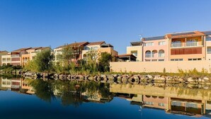 A waterfront property with a row of modern buildings, a calm lake reflecting the structures, and a stone wall in the foreground. - Studio Equipé 1.5km de la Plage (Le Barcarès)