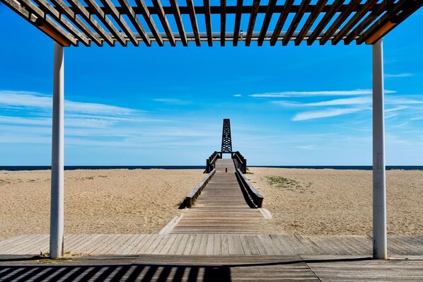 A wooden boardwalk leading to a pier with a lattice structure, surrounded by a sandy beach and clear blue sky.
