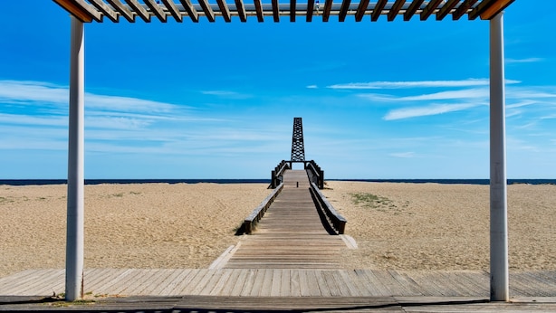 En træpromenade der fører til en mole med et gitterværk, omgivet af en sandstrand og klar blå himmel.