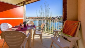 A balcony with white plastic chairs, a table with a purple tablecloth, and a view of a body of water and buildings in the distance. - Appart Abordable avec Terrasse Privée (Le Barcarès)