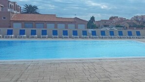 A swimming pool with blue water, surrounded by lounge chairs and a tiled deck. - Appart Abordable avec Terrasse Privée (Le Barcarès)