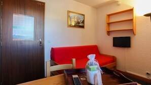 Een kleine kamer met een houten deur, een rode bank, een tafeltje met een afstandsbediening en een tas, en een aan de muur bevestigde televisie.