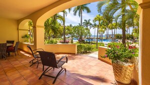 A covered patio with chairs and a table, overlooking a pool and palm trees.