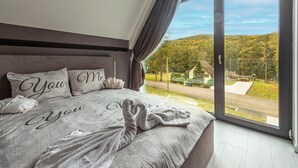 A bedroom with a large bed, a view of a green landscape through a sliding glass door, and a grey headboard.