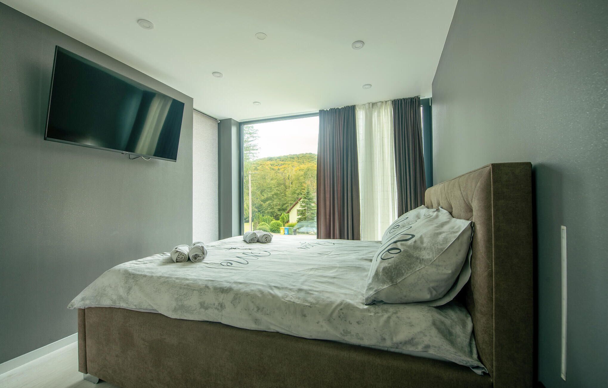 A bedroom with a bed, a TV mounted on the wall, and a view of a green landscape through the window.