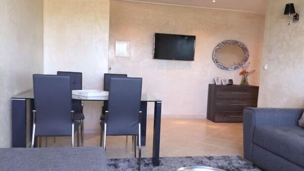 Un salon moderne avec un canapé gris, une table à manger avec des chaises, un téléviseur à écran plat fixé au mur et un miroir décoratif.