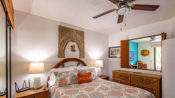 A bedroom with a bed, two bedside lamps, a wooden dresser, a ceiling fan, and a mirror. - Charming Retreat: 1-Bedroom Condo in Historic SJD (San José del Cabo)