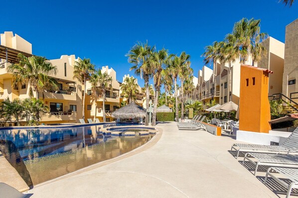 Een zwembadgedeelte van een resort met een helderblauwe lucht, palmbomen en moderne gebouwen met balkons.
