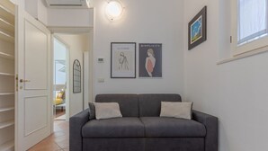 A grey sofa with two pillows, a white door, a framed artwork on the wall, and a window with blinds. - GuestHost - Manfredini Historical House in the heart of Ferrara (Ferrara)