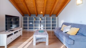 Een woonkamer met een houten plafond, een grijze bank, een witte salontafel en een boekenkast.