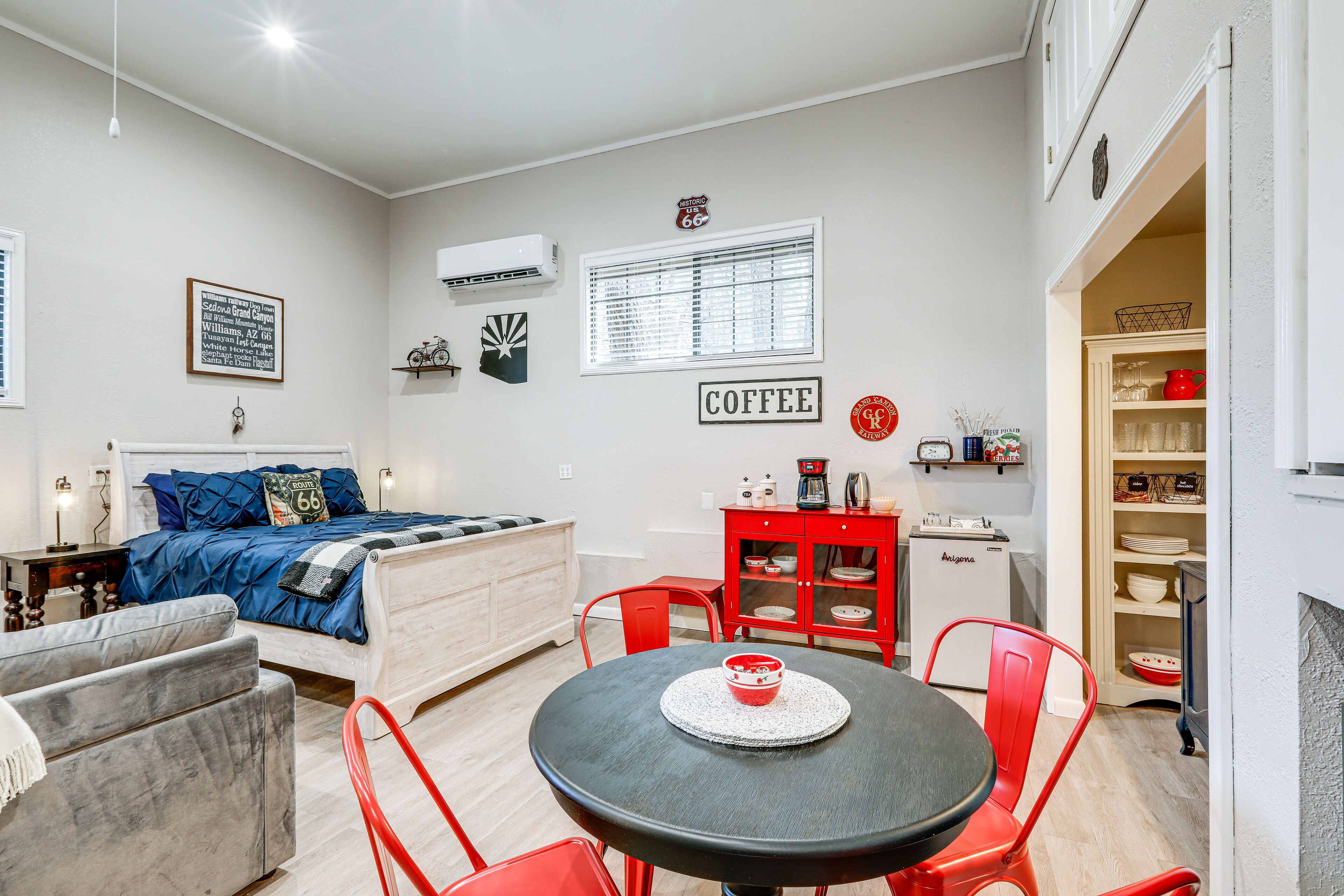 A compact living space with a bed, a dining table, red chairs, a small kitchenette, and a coffee sign on the wall.