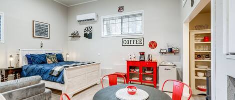 A compact living space with a bed, a dining table, red chairs, a small kitchenette, and a coffee sign on the wall.