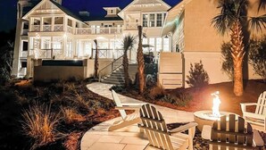 A large, multi-story house with a well-lit exterior, a visible staircase, outdoor seating area with chairs and a table, and a small fire feature. - Gulf Bridge by Stay on 30a (Inlet Beach)