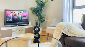 A living room with a flat-screen TV displaying a show, a potted plant, a clock, and a cozy seating area.