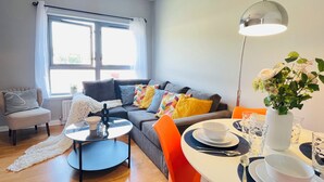 A modern living room with a grey sofa, colorful cushions, a coffee table, and a dining area with orange chairs and a white table.