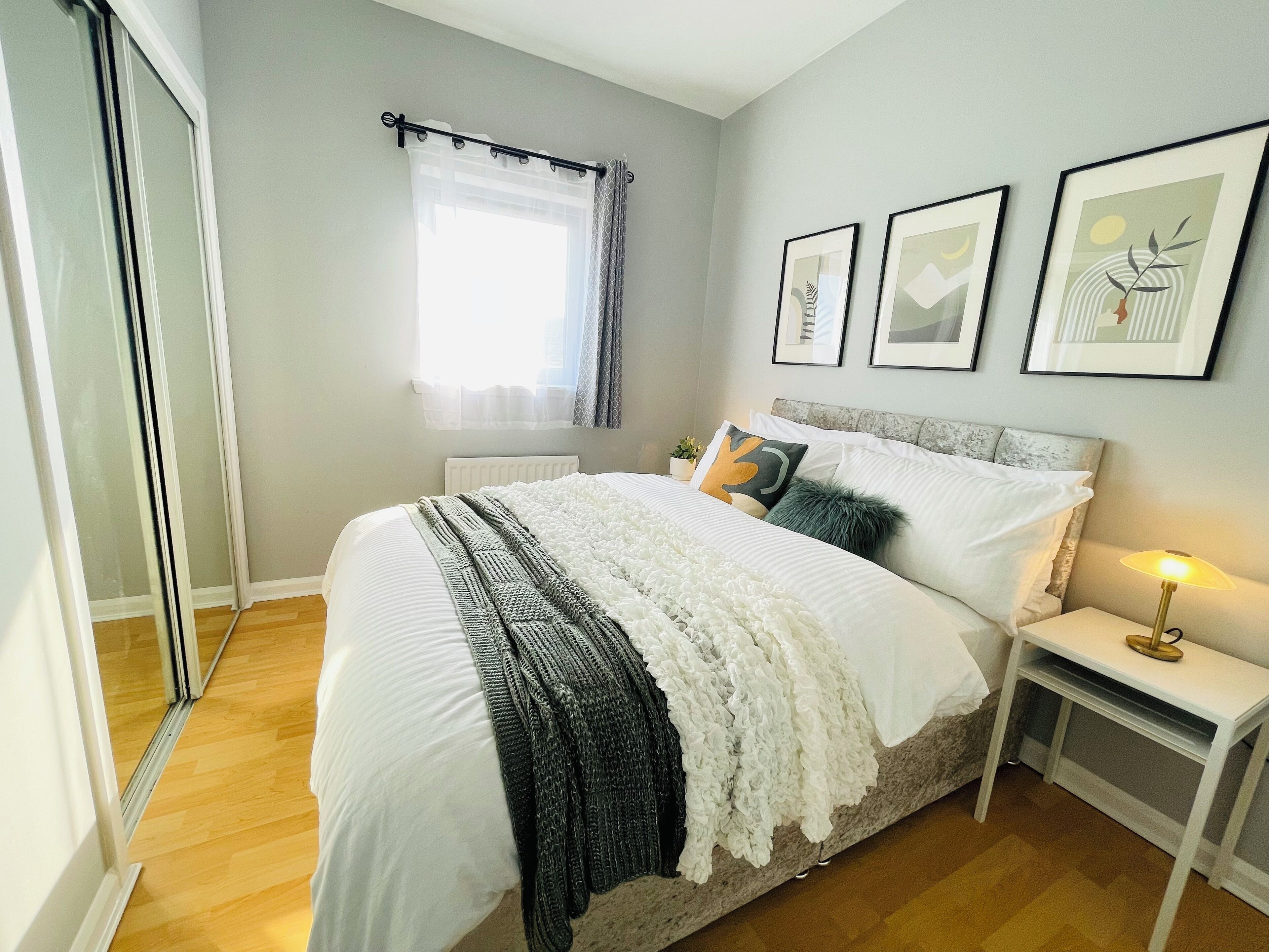 Un dormitorio con cama, mesita de noche, lámpara, ventana con cortinas y cuadros en la pared.