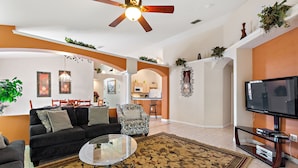 A living room with a sofa, coffee table, and TV. There is a kitchen in the background. - Soaring Eagle at Eagle Point by Shine Villas 102 (Kissimmee)