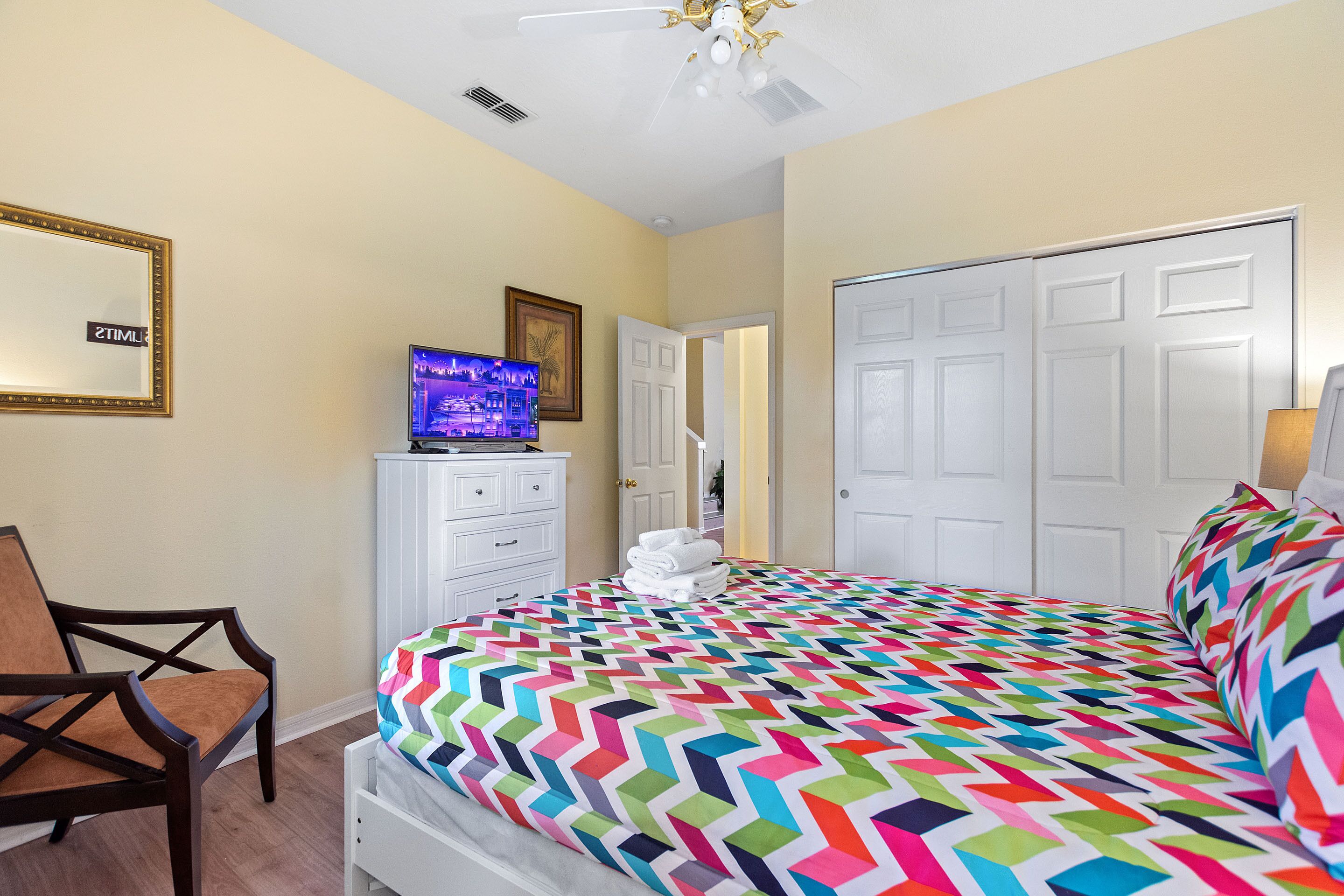 A bedroom with a colorful chevron-patterned bedspread, a television on a dresser, a chair, and a door leading to another room.
