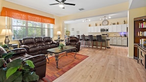 A spacious living room with leather sofas, a coffee table, and a dining area with chairs. A kitchen with white cabinets and a clock is visible in the background. - Greenview Bliss by Shine Villas #031 (Davenport)