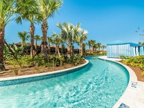 A swimming pool with a curved edge, surrounded by palm trees and a clear blue sky. - Summer Solstice @ Windsor Westside by Shine Villas #114 (Kissimmee)