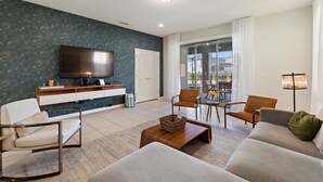A modern living room with a flat-screen TV, a sofa, two armchairs, a coffee table, and a sliding glass door leading to a balcony. - Summer Solstice @ Windsor Westside by Shine Villas #114 (Kissimmee)