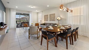 A modern dining area with a round table, chairs, and a chandelier. There is a sofa and a coffee table in the background. - Summer Solstice @ Windsor Westside by Shine Villas #114 (Kissimmee)