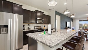 A modern kitchen with dark cabinets, stainless steel appliances, a granite countertop, and a bar area with stools. - Summer Solstice @ Windsor Westside by Shine Villas #114 (Kissimmee)