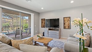 A modern living room with a sofa, coffee table, and TV. There is a sliding glass door leading to an outdoor patio with a pool and chairs. - Blissful Haven @ Windsor Westside by Shine Villas 995 (Kissimmee)