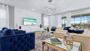 A modern living room with a beige sofa, blue armchairs, and a glass coffee table. A large TV screen displays "SHINE villas".