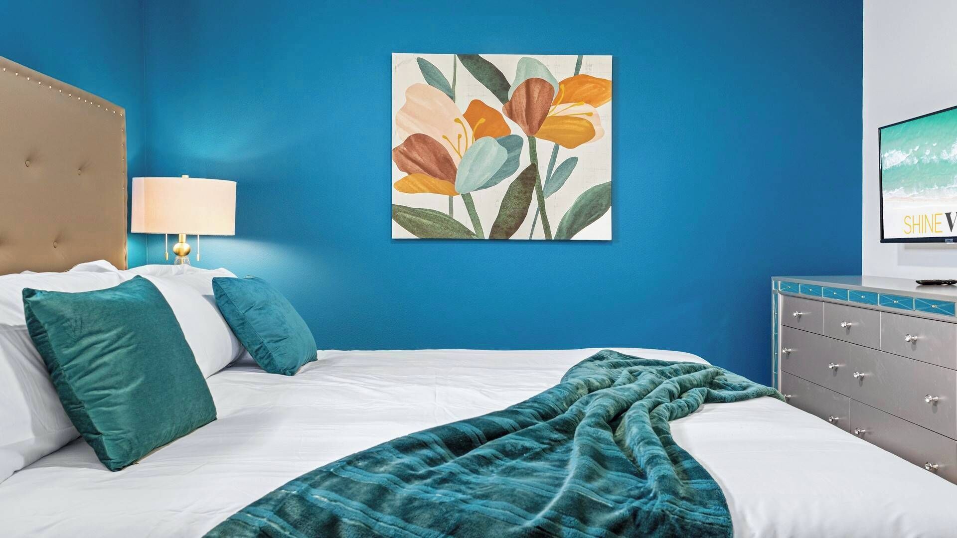 Une chambre à coucher avec un lit, des coussins et une couverture de couleur turquoise, une tête de lit, une table de chevet avec une lampe, un téléviseur fixé au mur et un tableau représentant des fleurs.