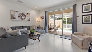 Amplia sala de estar con un sofá gris, un sillón beige y una mesa de centro. Hay un ventanal corredizo que da a un patio exterior con piscina.