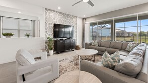 Un salon moderne avec un canapé d’angle, un fauteuil blanc, une table basse et un téléviseur à écran plat fixé au mur, lequel est décoré d’un motif géométrique.